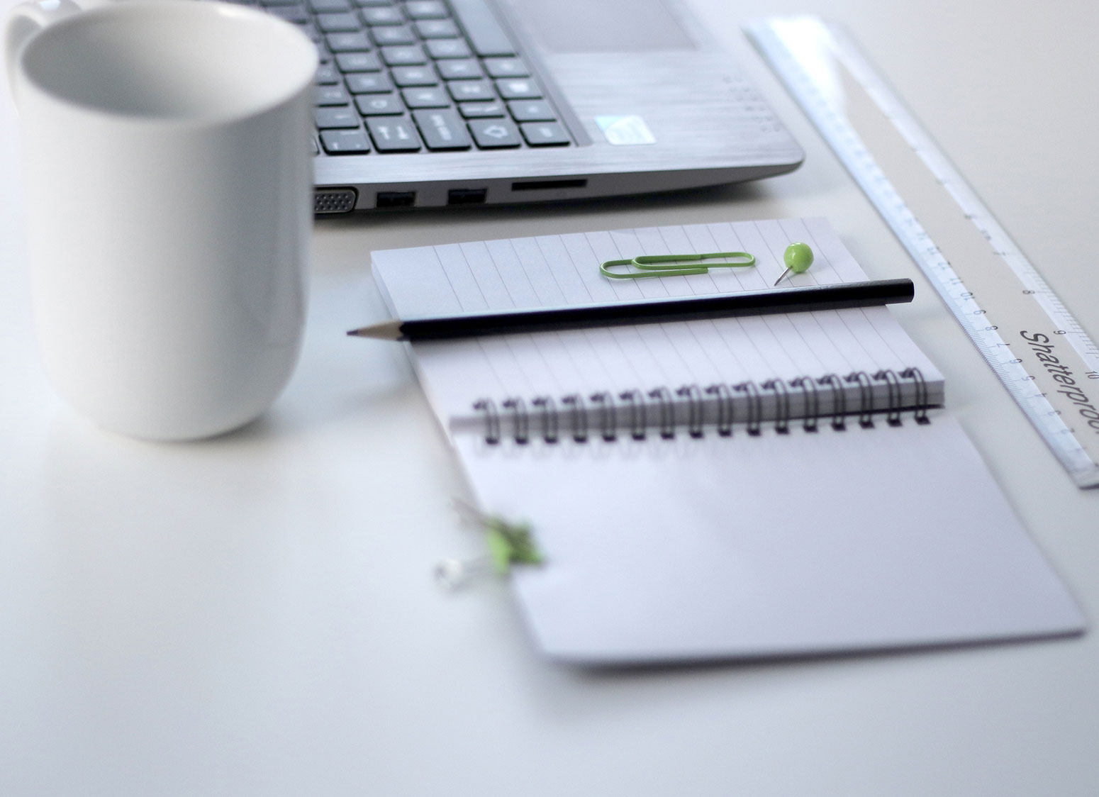 blank notebook, paper clip, coffee cup and laptop on white desk